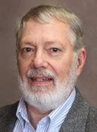 A man with gray hair and a beard is wearing a gray suit jacket over a blue and white striped shirt. He is posing in front of a neutral background.